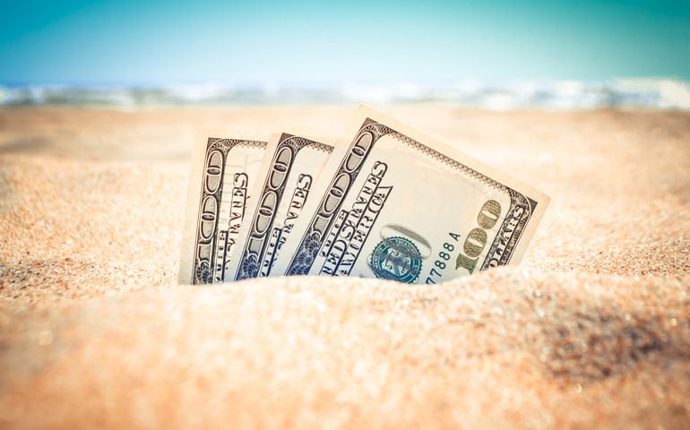 Money half covered with sand lie on sandy beach near sea ocean waves on sunny summer day close-up. Money grows out of the ground. Concept finance money holiday relax vacation.