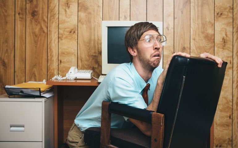 A terrified man and office in 1980's - 1990's style, complete with vintage computer and technology of the time, hides behind his chair at his desk with a look of fear on his face at some crisis or disaster occurring at work. Wood paneling on the wall in the background. Horizontal with copy space.