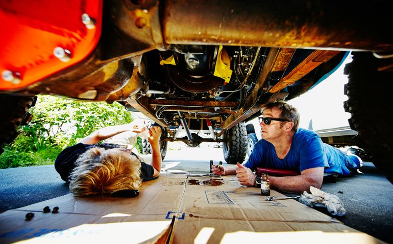 Father and son lying on driveway under off road vehicle replacing oil pan gasket