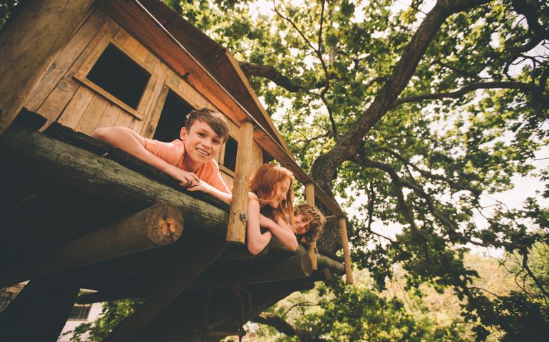 Portrait of a boy smiling at the camera while playing with two friends in rustic wooden treehouse in the branches of a big tree with healthy green leaves