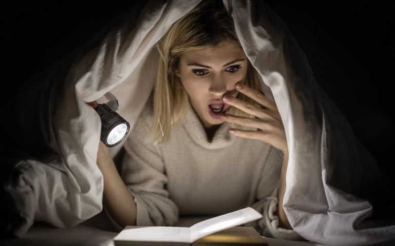 Beautiful young woman reading book under bed cover and holding a flashlight.
