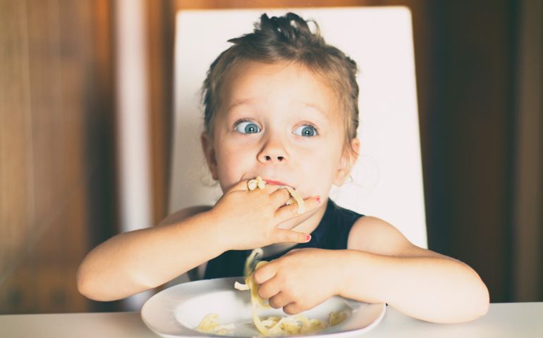 Three years old girl sitting at the table in the kitchen and eating pasta.