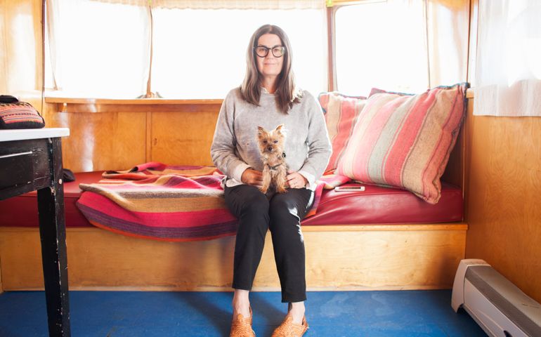 A woman wearing glasses and a slight smile with her small dog on her lap looking at the camera
