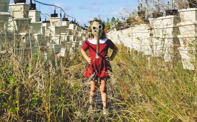 Young child in a vintage looking photograph stands in an empty field of overgrown weeds and empty vertical gardens wearing a gas mask. Commentary on pollution, pesticides, and a dystopian future