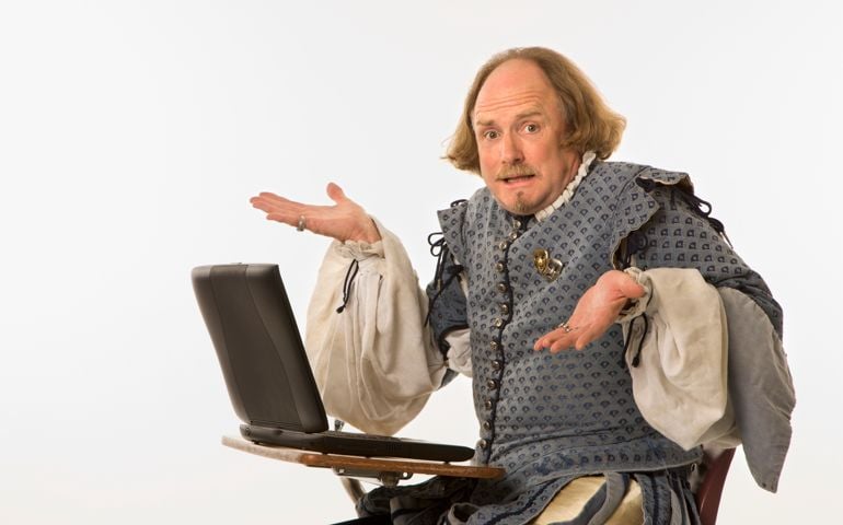 William Shakespeare in period clothing sitting in school desk with laptop computer shrugging at viewer.