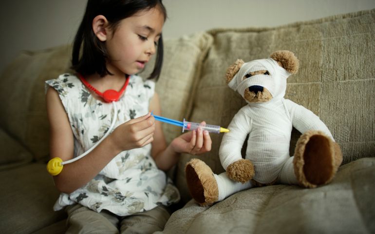 Girl plays doctor doctor giving bandaged teddy an injection with a toy syringe.