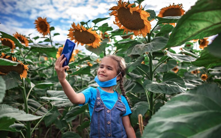 Local travel and new normal. Happy kid in medical mask making selfie with smartphone in the sunflower field during family car trip, summer outdoor lifestyle