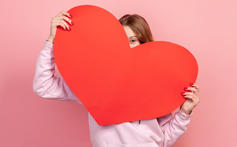 Portrait of playful young brunette female in hoodie hiding behind red paper heart, demonstrating love, present for valentines day. Indoor studio shot isolated on pink background