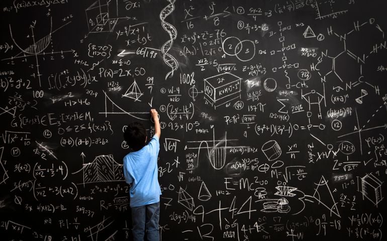 A young boy stands drawing on a huge chalkboard filled with mathematical equations
