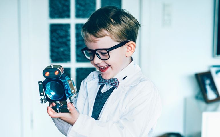 Boy dressed as a professor plays with a toy robot. The boy wears a lab coat, glasses and a bowtie. The boy smiles.