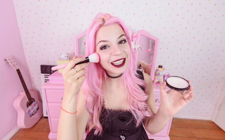 Woman with dyed hair in bedroom holding make up brush and putting on cosmetics, smiling with cheerful expression
