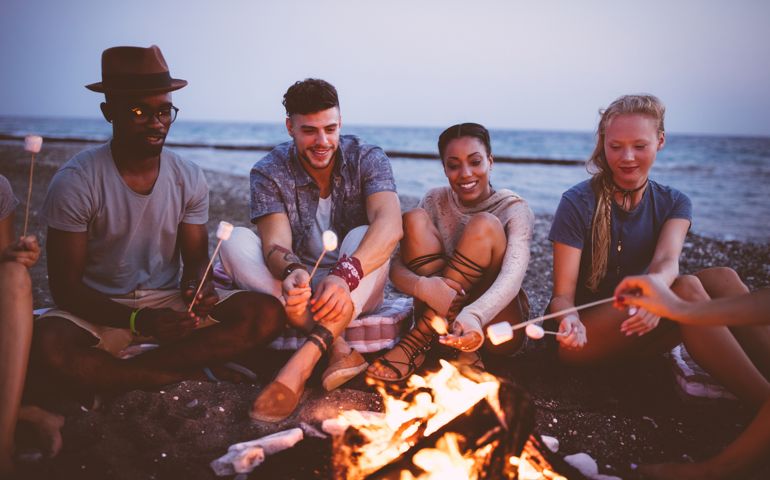 Young multi-ethnic hipster friends on summer holidays roasting marshmallows over bonfire at the beach