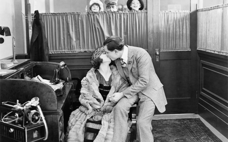 Couple kissing in an office while children and a messenger are peeking over a curtain