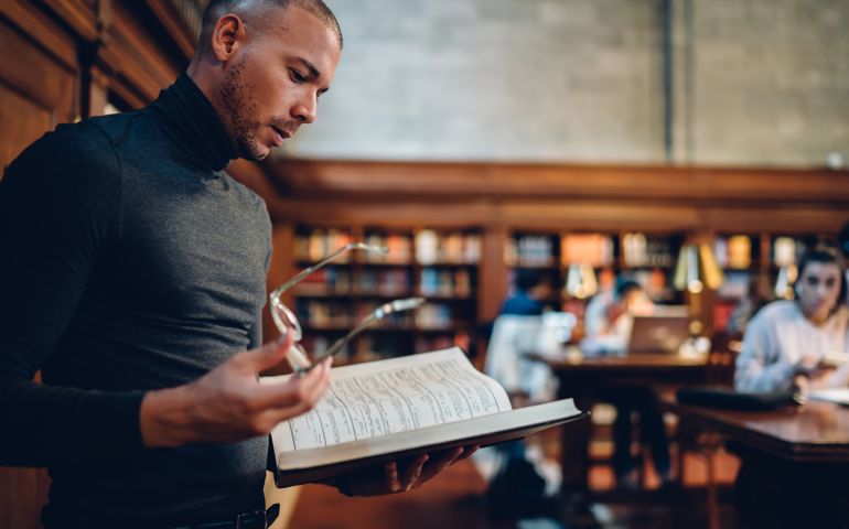 Smart intellectual male lawyer reading books making researcher for work while standing in public library, pensive concentrated journalist analyzing interesting information from literature manuals