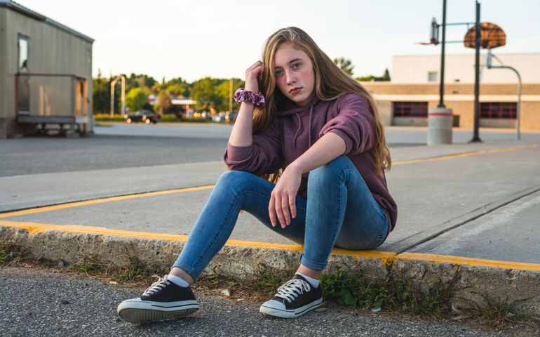 Sad teen girl sitting on a curb in front of a school.
