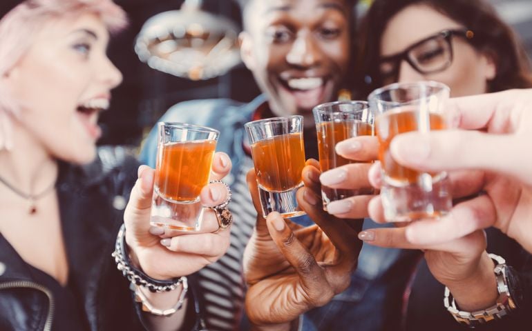 Multi ethnic group of happy friends - caucasian and afro american - drinking shots in the pub. Focus on hands and shot glasses.