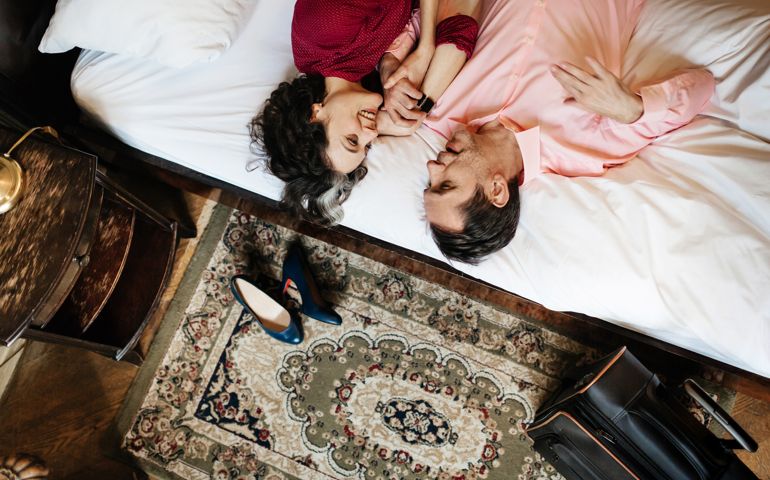 A mature, loving couple is looking at each other lying on the white sheets of a hotel bed. They are holding hands and seem to be in love. On the floor, there's an old carpet, a suitcase and the woman's shoes.