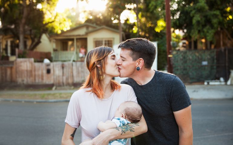 A husband kissing his wife who is holding their newborn baby on a suburban street