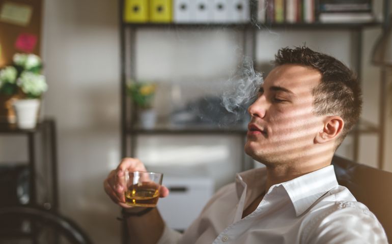 Handsome looking businessman in white shirt, sitting in his office after work, enjoying a cigarette.