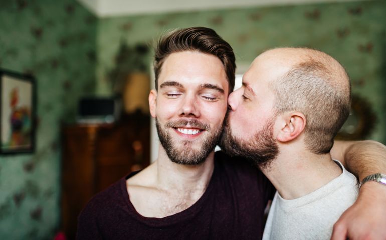 A gay man kissing his gay partner, who is smiling, on the cheek.