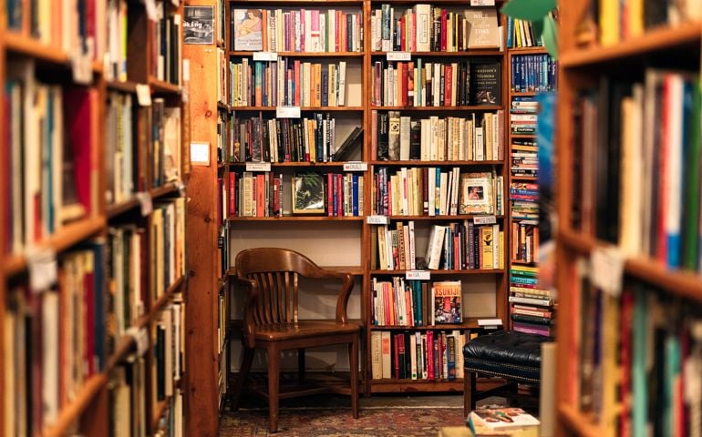 An array of book titles under a variety of category headings on bookshelves above a wooden chair in the corner of a San Francisco second-hand bookstore.
