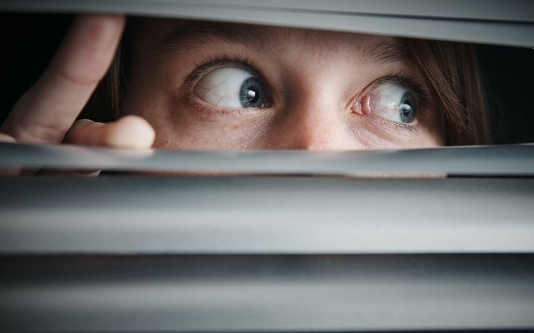 A young woman looks to the side through blinds, terrified.