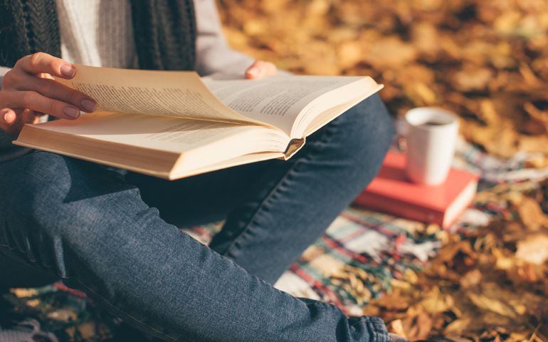 Cropped image of young woman sitting on blanket, reading book and drinking coffee or tea in autumn garden