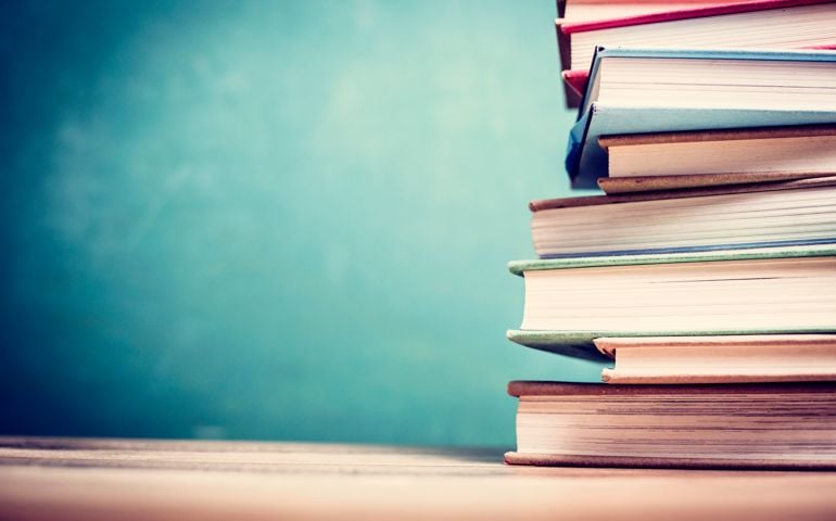 Textbooks stacked on wooden school desk in front of green chalkboard.
