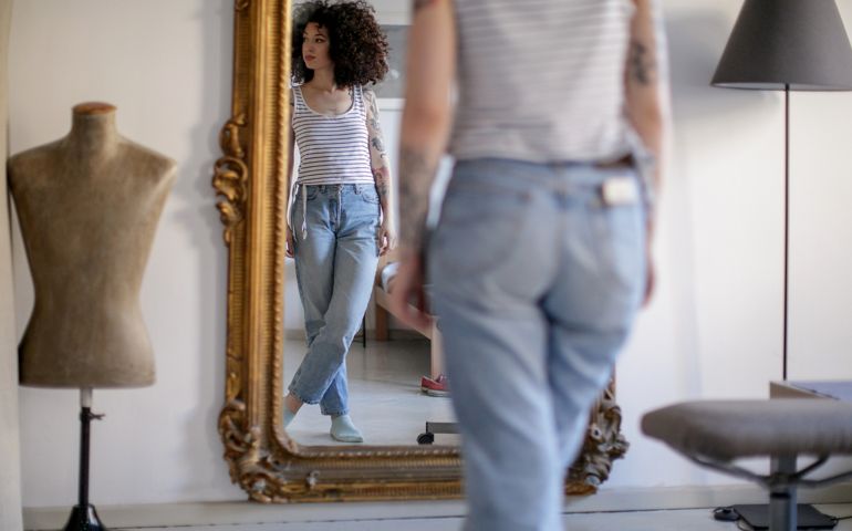 Reflection of a young tattooed woman with brown curly hair standing in front of the mirror and looking away