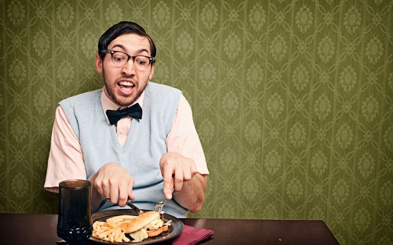 A nerdy young man eats an unhealthy dinner of hamburger and fries in his vintage decorated living room