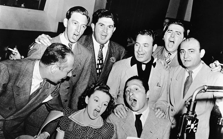 American actors Bert Lahr (1895 - 1967) (far right), Ray Bolger (1904 - 1987) (back row, right), Judy Garland (1922 - 1969) (sitting, right), composer Harold Arlen (1905 - 1986) (sitting left), and various MGM and music publishing executives sing songs from the 1939 film musical 'The Wizard of Oz' around a microphone in the NBC radio studio, circa 1939.