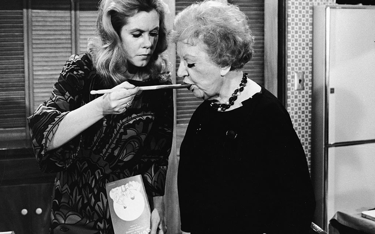 American actress Elizabeth Montgomery (1933 - 1995) (left, as Samantha Stephens) feeds American actress Marion Lorne (1883 - 1968) (as Aunt Clara) with a wooden cooking spoon as she holds a cookbook entitled 'Sukiyaki: the art of Japanese cooking' in a still from the episode 'A Majority of Two' of the television sitcom 'Bewitched,' April 11, 1968.