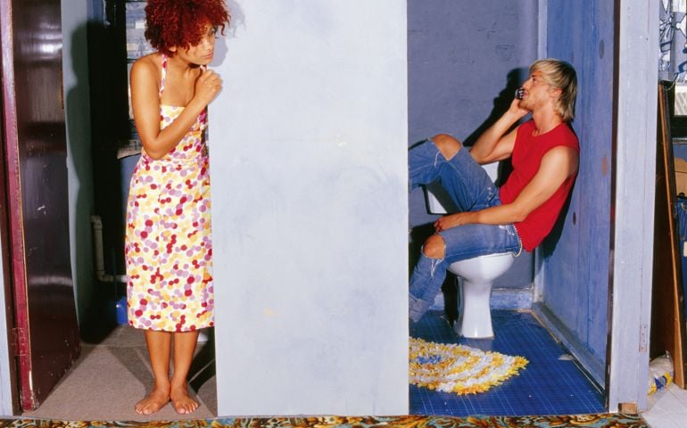 Young man on toilet using mobile phone, woman looking around the corner