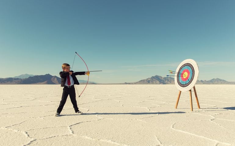 A young boy dressed in business suit, glasses and tie holds shoots a bow and arrow towards a target on the Bonneville Salt Flats in Utah. His business has found success and it mark for the future. He is smiling at the camera in front of blue skies.