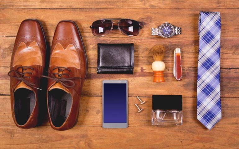Variety of men's accessories organized in knolling arrangement. Items include: dress shoes, tie, sunglasses, wallet, watch, shaving brush, cologne, and smart phone. The items all lie on a wooden desk or table. Men's personal accessories, clothing themes. Father's Day. Fashion. Business. Retail.