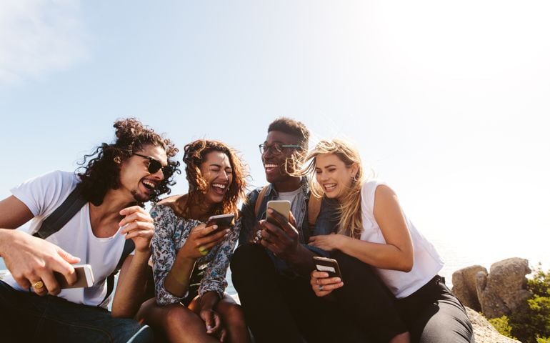 Young group of people sitting on top of mountain using smart phones and smiling. Diverse friends enjoying a day out.