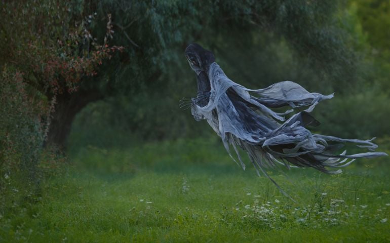 Handmade Death flies through the air with a wavy mantle into the forest above the lawn with green grass.