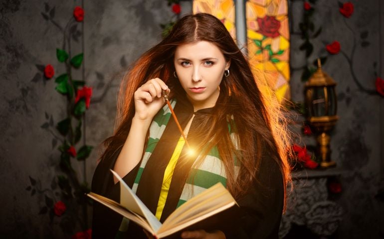 A young woman in a black mantle with a striped scarf around her neck casts a spell from a book and conjures with a wand of magic.