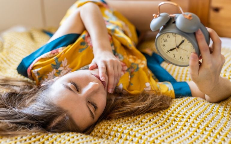 Beautiful young woman of 30 in bed, late in the morning, surprized by the alarm clock time, oversleeping and too late for work or school