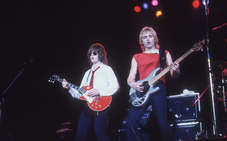 circa 1982: American rock guitarist Elliot Easton (left) and bassist Benjamin Orr performing on stage with the group the Cars. Easton wears sunglasses, a white shirt, and a red tie. Orr wears a red T-shirt with an asymmetrical neckline and black jeans. Both have shag hairstyles.