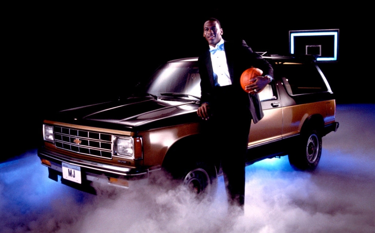 Portrait of professional basketball player Michael Jordan, of the Chicago Bulls, dressed in a tuxedo with a basketball under one arm as he poses with Chevrolet Suburban automobile, Chicago, Illinois, 1986.