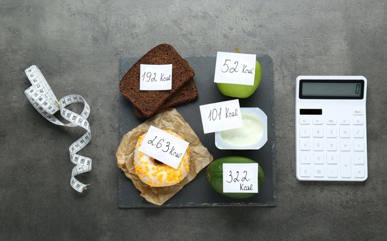Calculator and food products with calorific value tags on dark grey table, flat lay. Weight loss concept