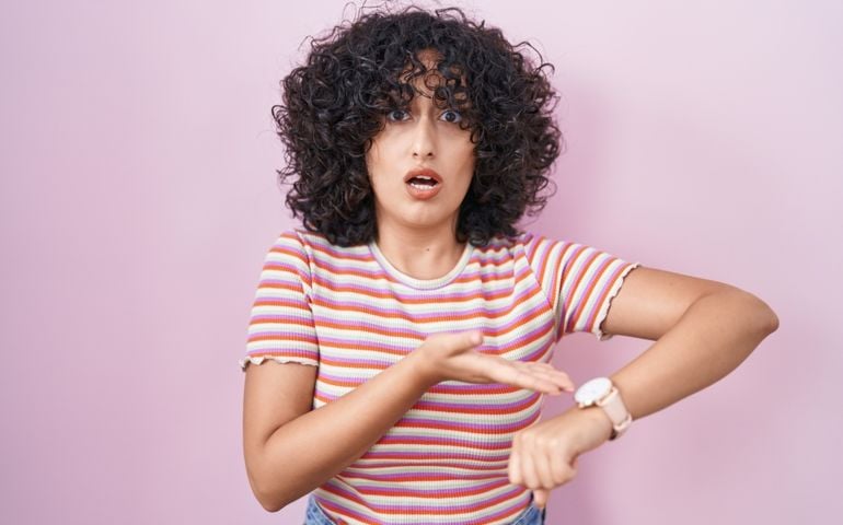 Young middle east woman standing over pink background in hurry pointing to watch time, impatience