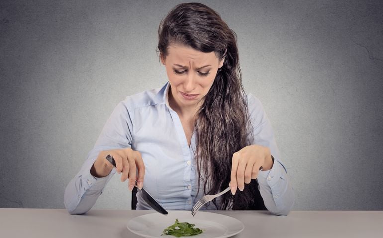 Young woman tired of diet restrictions eating green salad sitting at table isolated grey wall background. Human face expression emotion. Nutrition concept