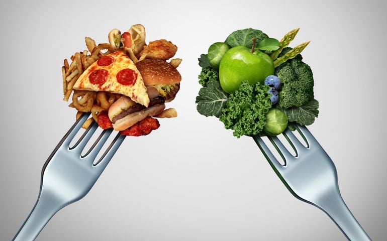 Diet struggle and decision concept and nutrition choices dilemma between healthy good fresh fruit and vegetables or cholesterol rich fast food with two dinner forks competing to decide what to eat.