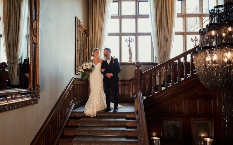 Bride linking arms with her father as they walk down the stairs on her wedding day. He is leaning over to kiss her cheek as she smiles, holding a bouquet of flowers.