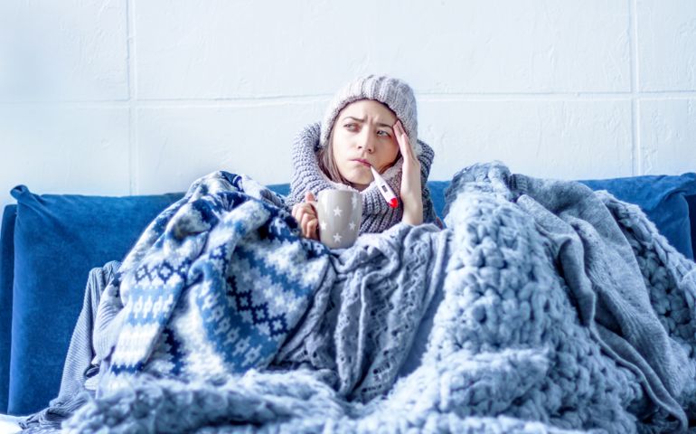 Sick exhausted girl in white warm hat wrapped herself in scarves and blankets is sitting in bed. Young woman with fever is drinking tea and measuring temperature with thermometer. Winter cold and flu.
