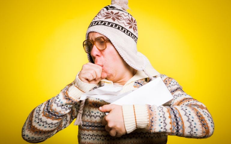 Funny stock image of sick young man with cold and flu. Studio shot on a yellow background, this young man is blowing his nose and coughing violently into his fist, and is wearing a winter sweater and hat. Winter time is cold and many people get sick during the colder months.