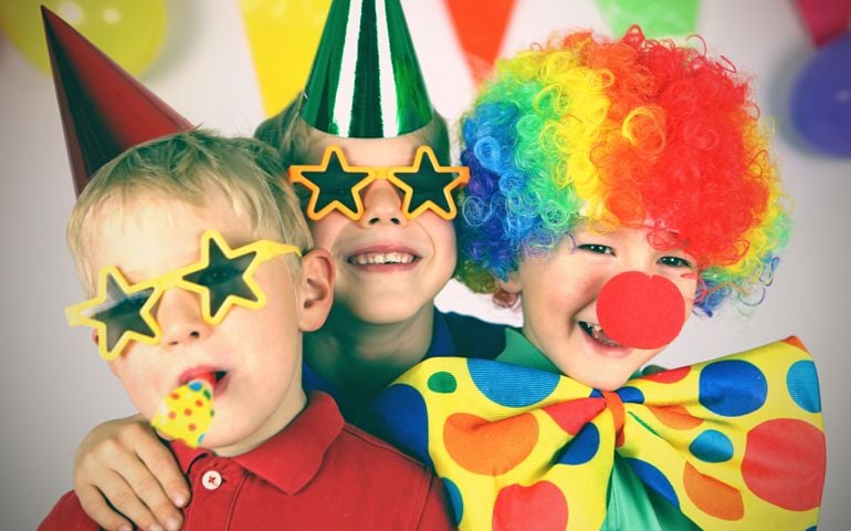 Three boys dressed up in colorful party clothing are celebrating a birthday. One child is wearing a colorful clown wig, a red nose, and a giant bow tie, the other two boys have party hats and funny sunglasses on, one of them is blowing a party blower. This is a tinted, retro style image of the childrens party.