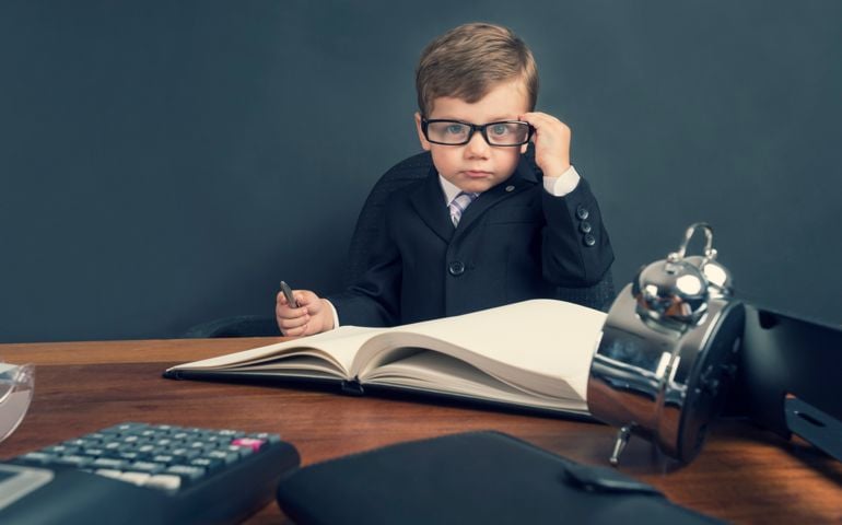 Young boy dressed in a suit working at a large desk. The desk has a calculator, Filofax, phone and clock. He is very busy and there is a lot of work on his desk. He is wearing glasses and looks like an accountant.
